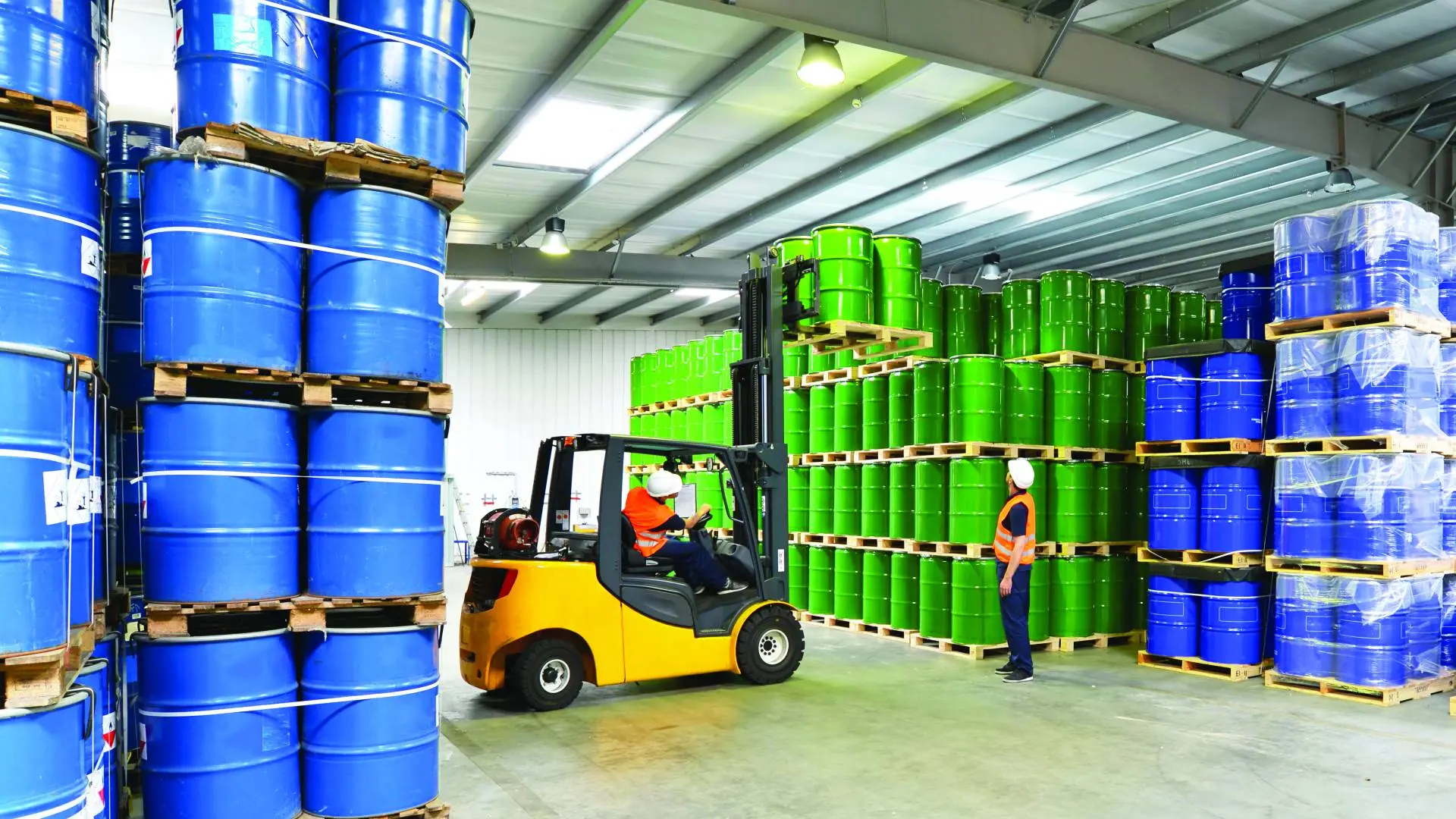 Depoda mavi ve yeşil varillerin arasında çalışan forklift operatörü